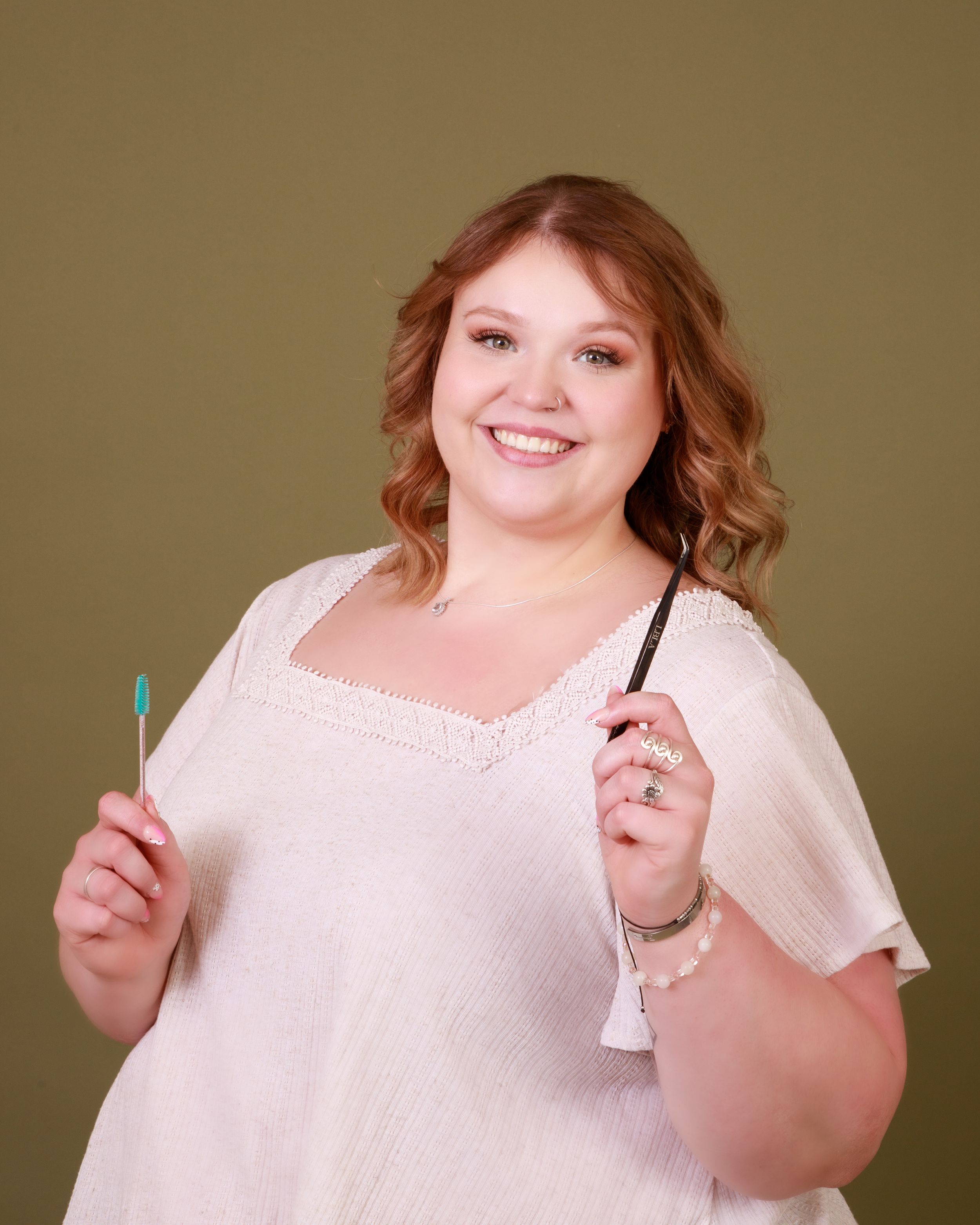 A person in a white lace top smiles while holding beauty products against an olive background.