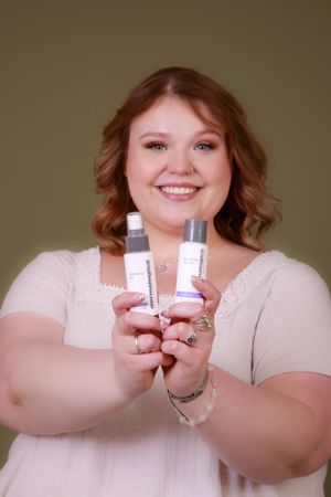 A person displays beauty products while wearing a white lace top against an olive background.