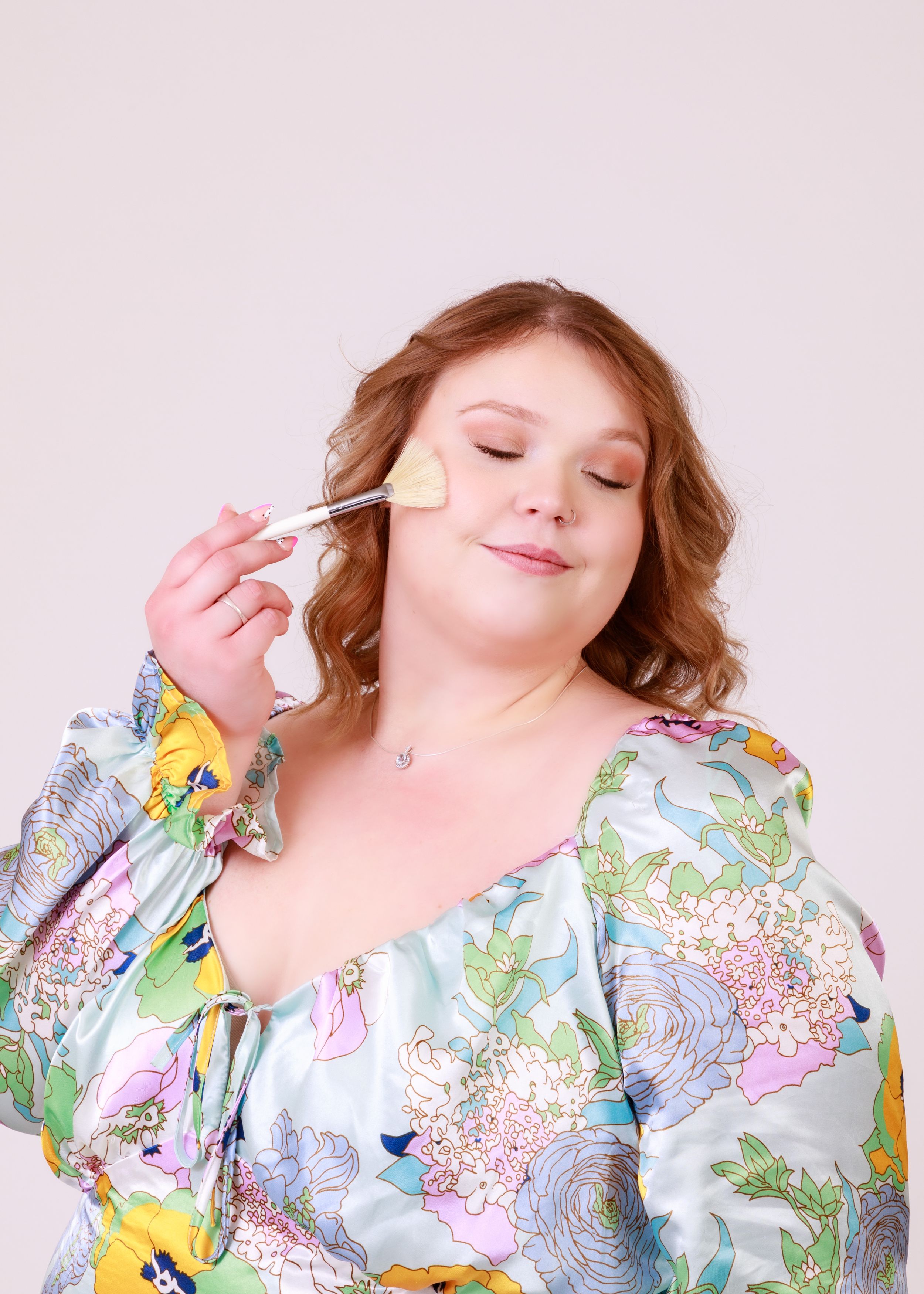 A person in a floral print top applies makeup with a brush while posing with eyes closed.