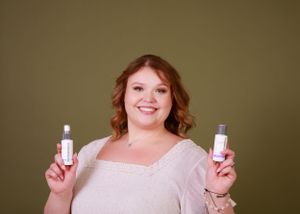 A person holds up beauty products while wearing white against an olive background.