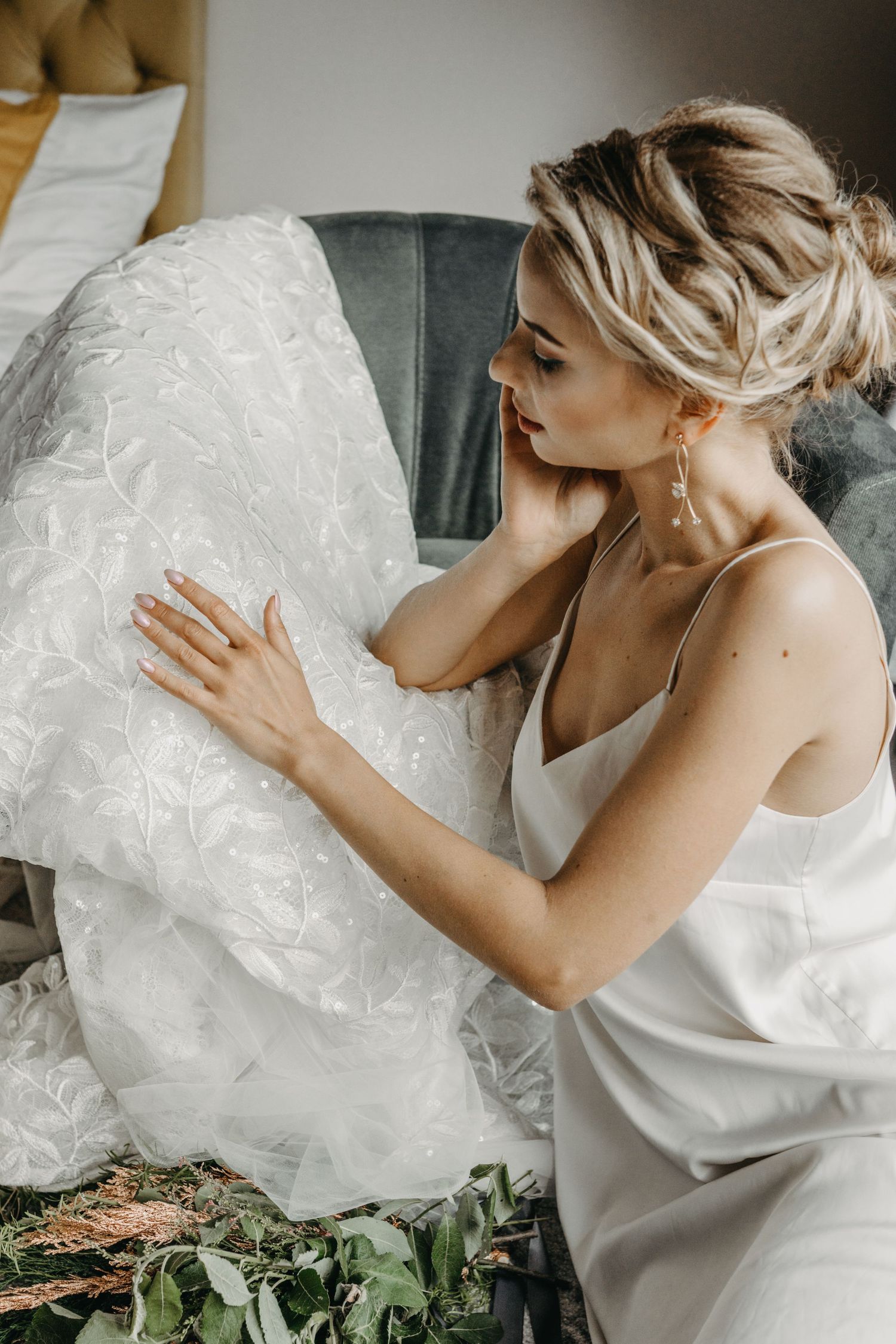 Blonde bride in silk slip dress admires her lace wedding gown while seated, wearing elegant drop earrings.