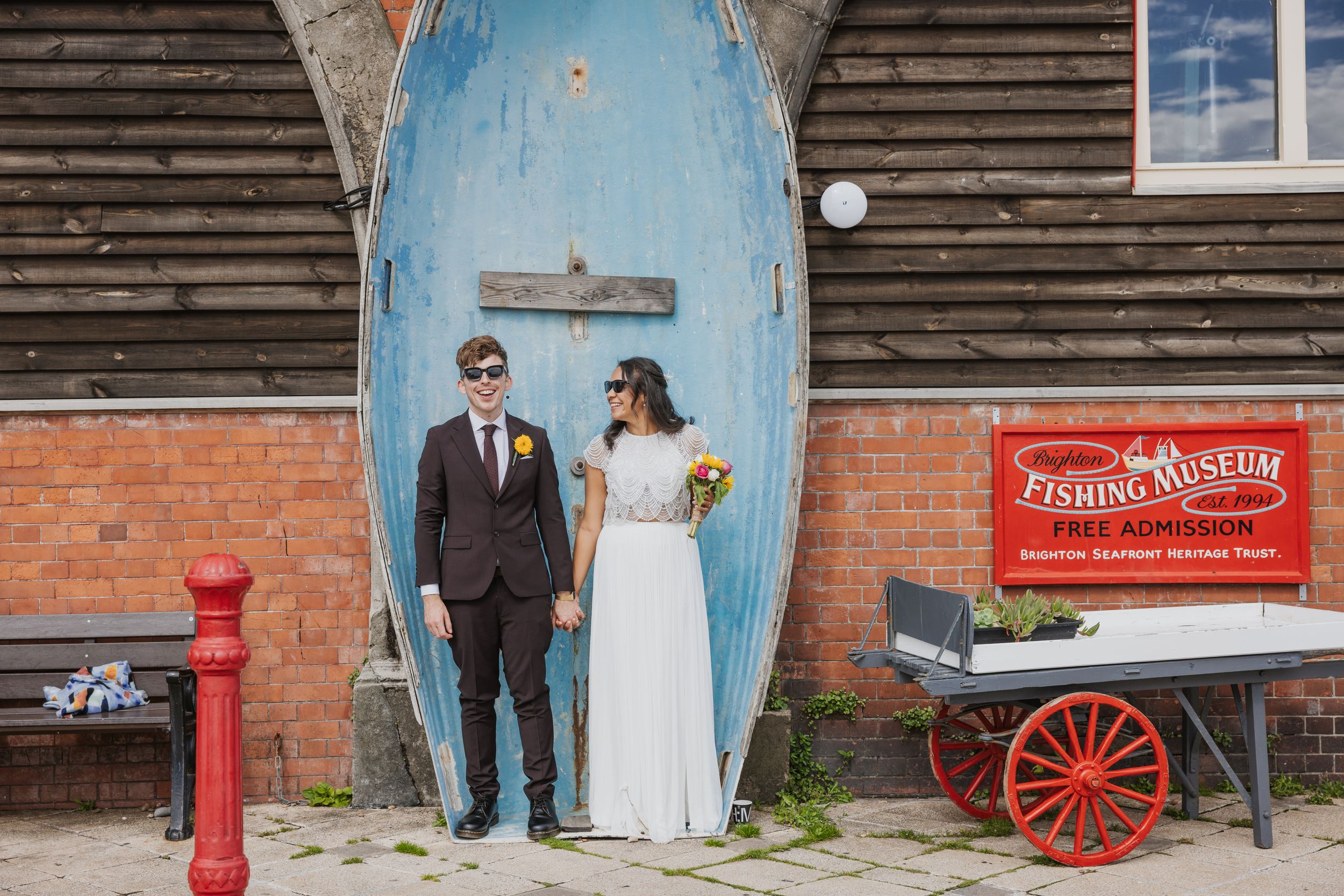 a couple standing in front of a boat on Brighton seafront holding hands wearing wedding dress and suit