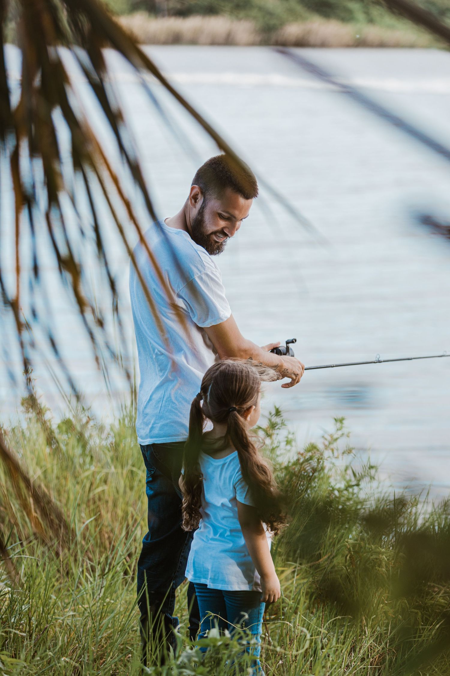 Parent and child fishing together near palm trees at waters edge.