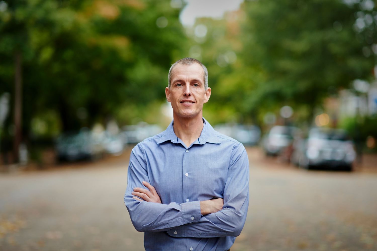 Professional outdoor portrait of person in light blue shirt against blurred tree background.