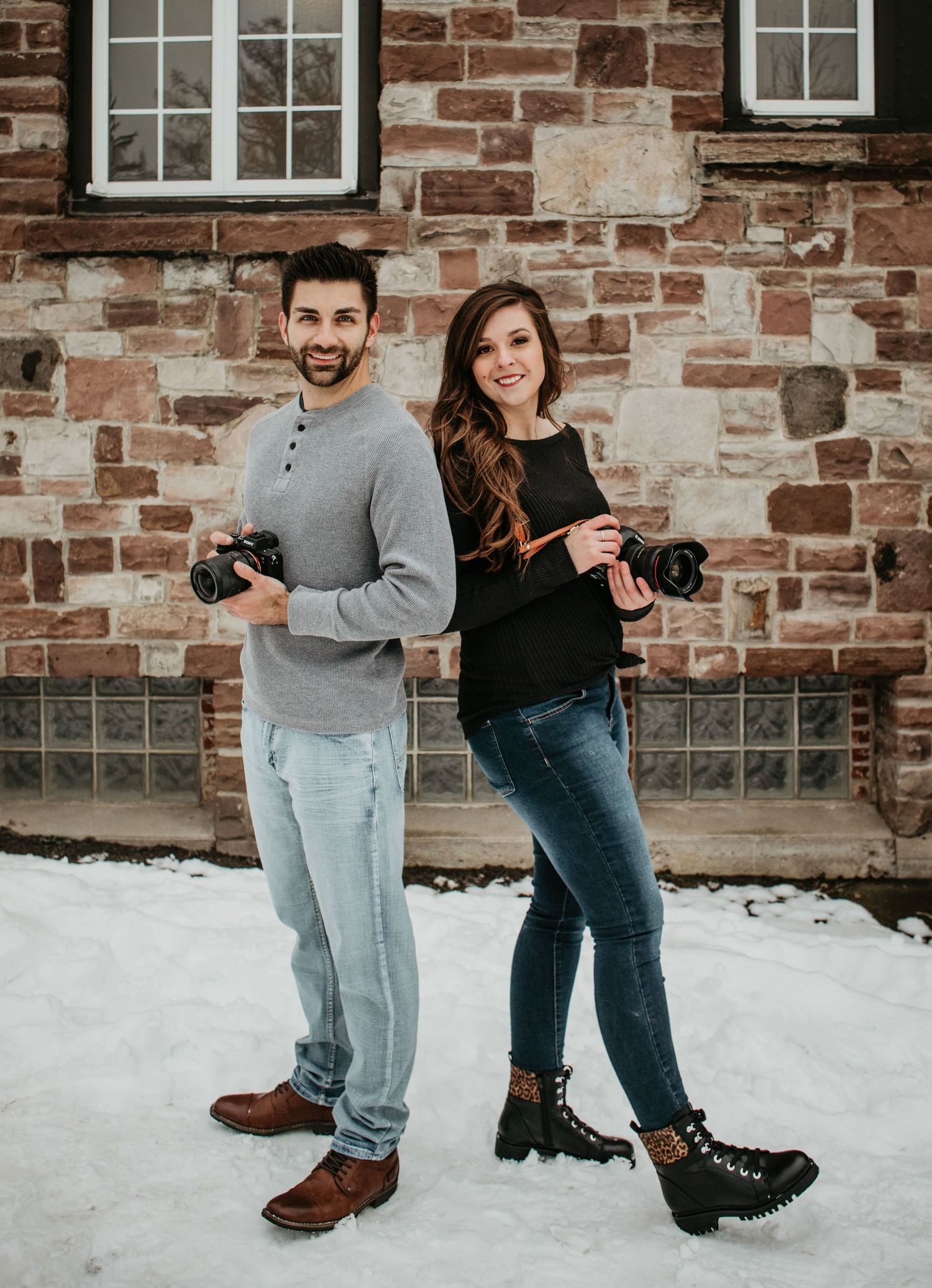 Two photographers with cameras standing against rustic brick wall in winter setting.