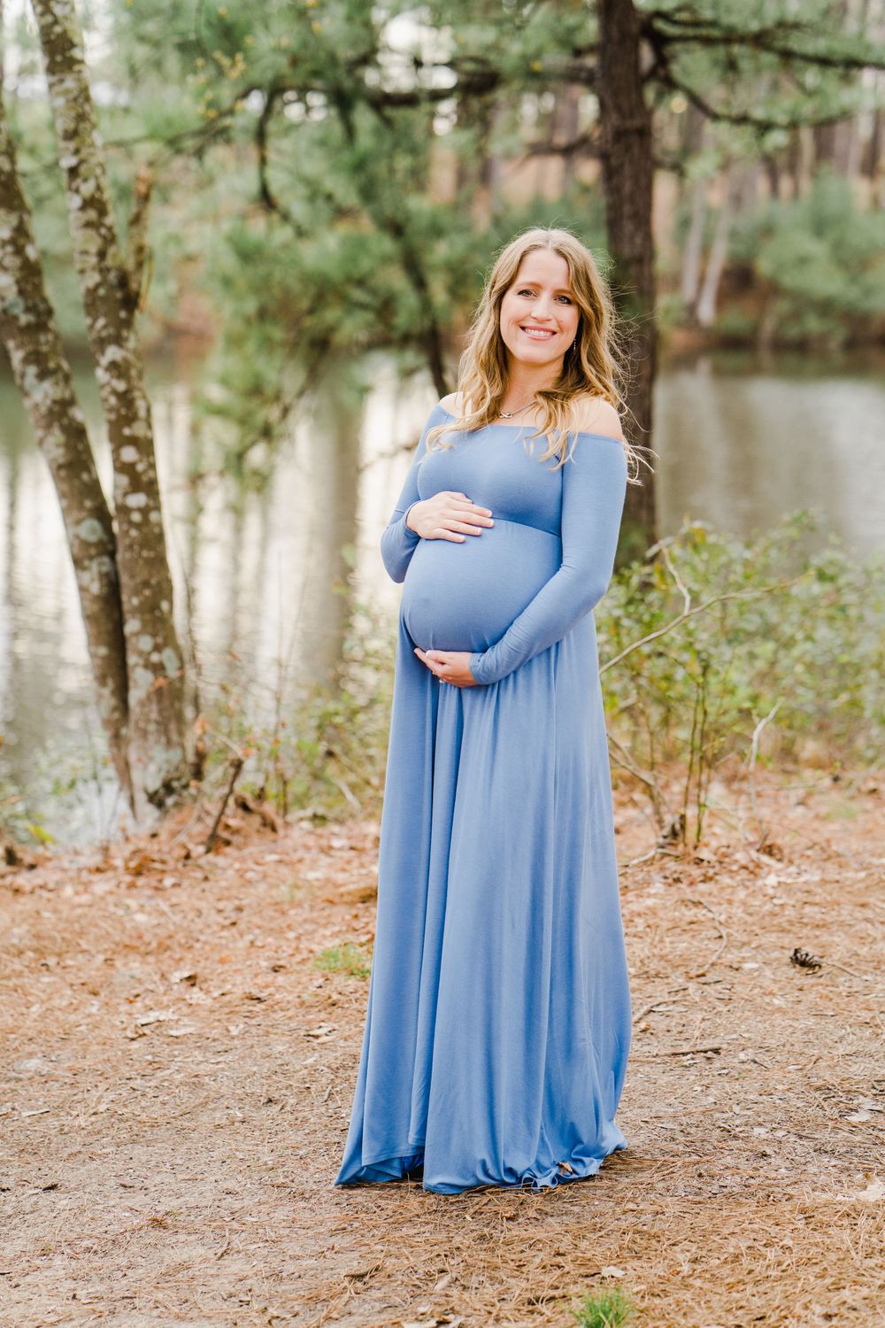 Expectant mother in long blue off-shoulder dress stands smiling by a lake surrounded by pine trees.