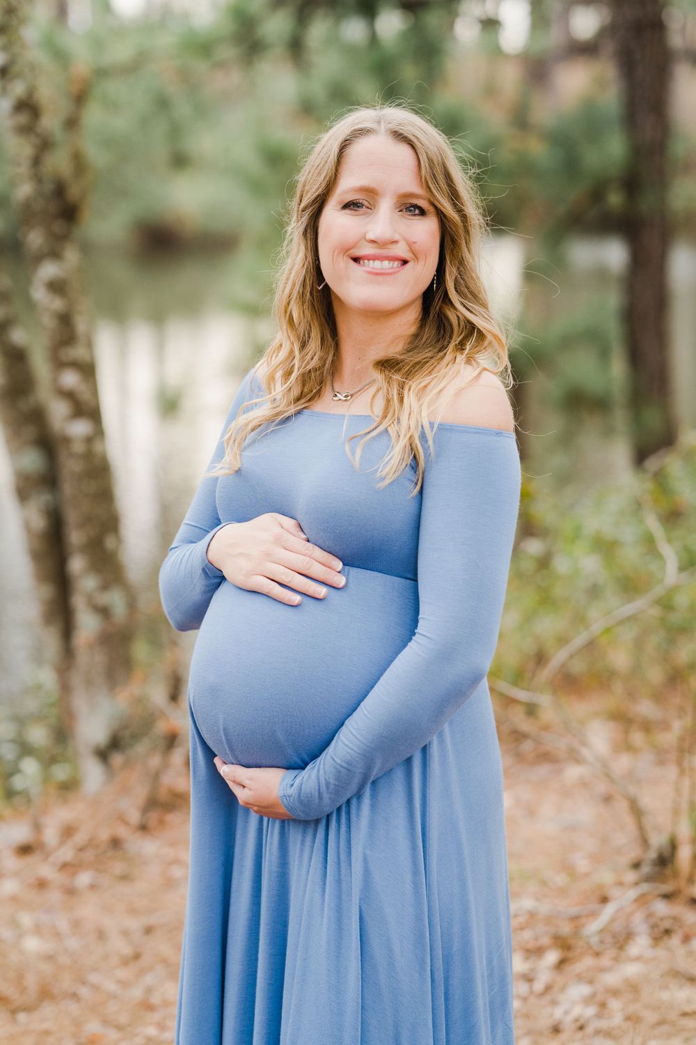 Pregnant woman in long blue dress smiles while cradling her baby bump outdoors.