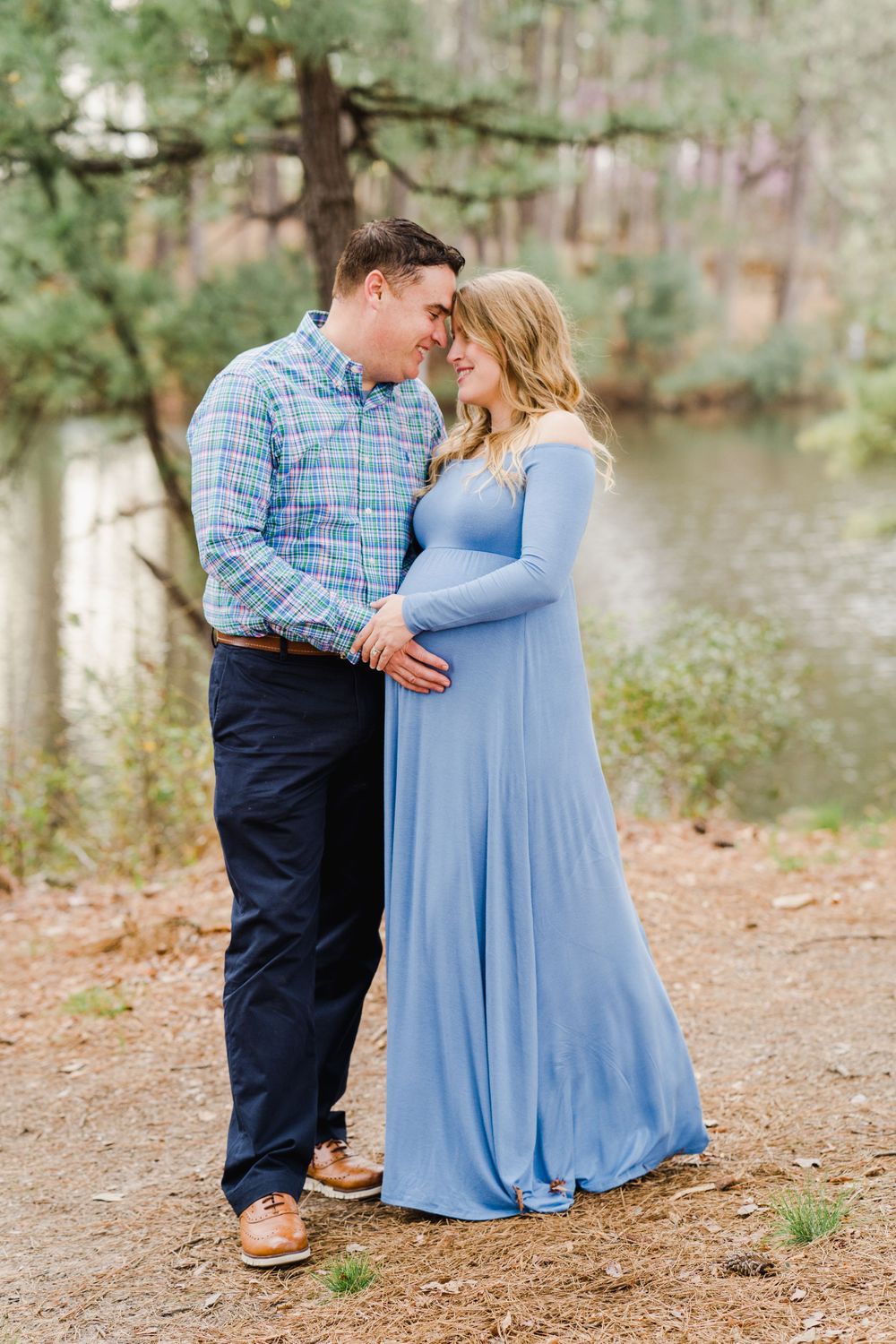 Couple shares intimate moment during maternity photoshoot by lake with pine trees in background.