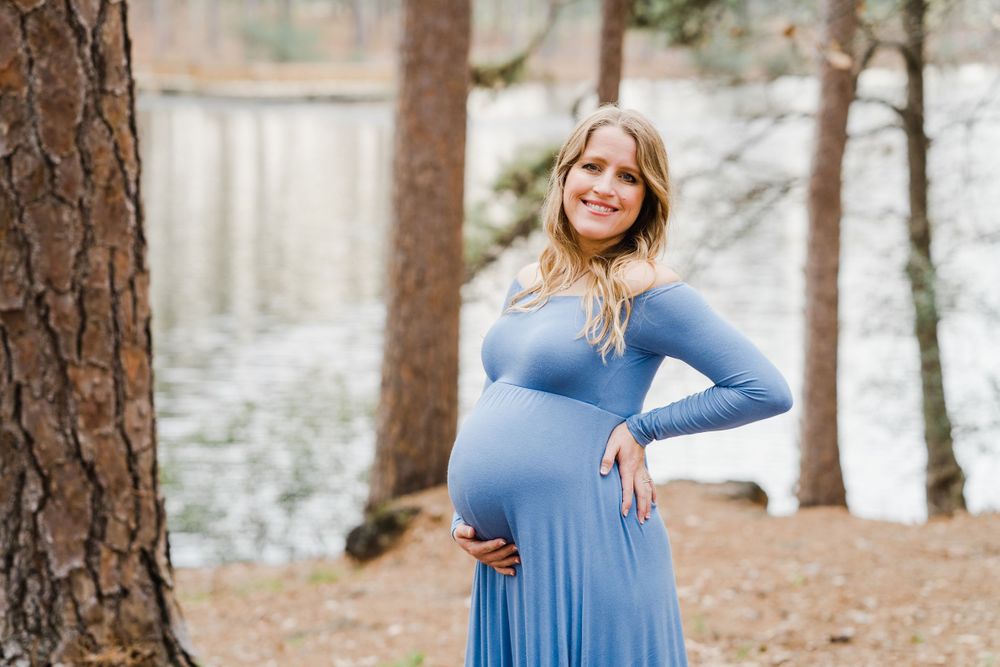Woman in blue maxi dress poses during maternity shoot in pine forest setting.