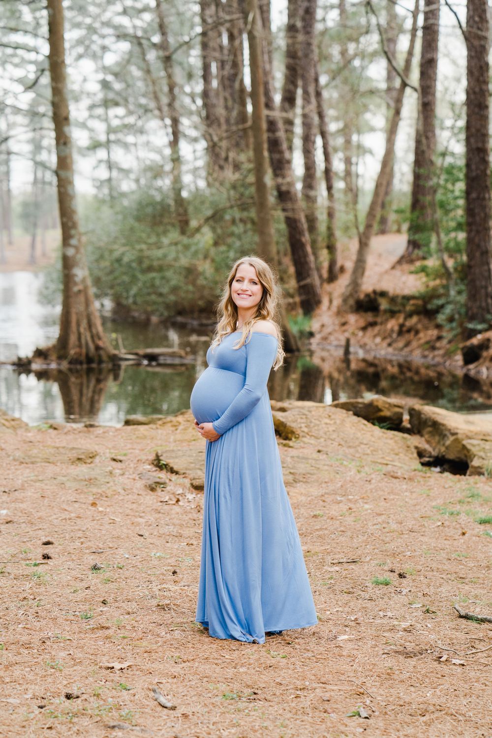 Expectant mother in flowing blue dress poses by lake during outdoor maternity session.