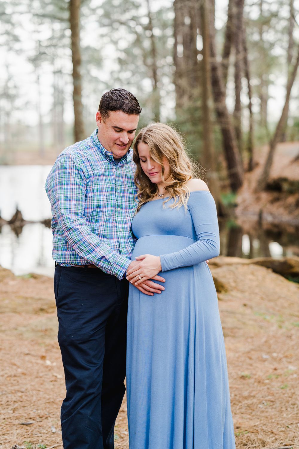 A couple in coordinating blue outfits embrace during a maternity photoshoot by a lake in a wooded setting.