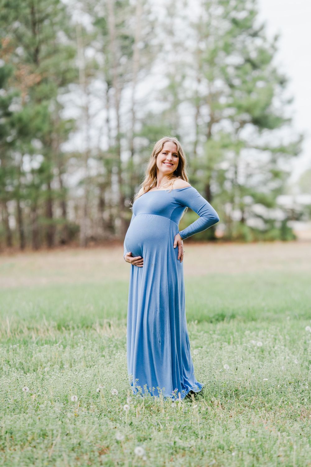 Expectant mother in a long blue dress poses in an open field surrounded by pine trees.