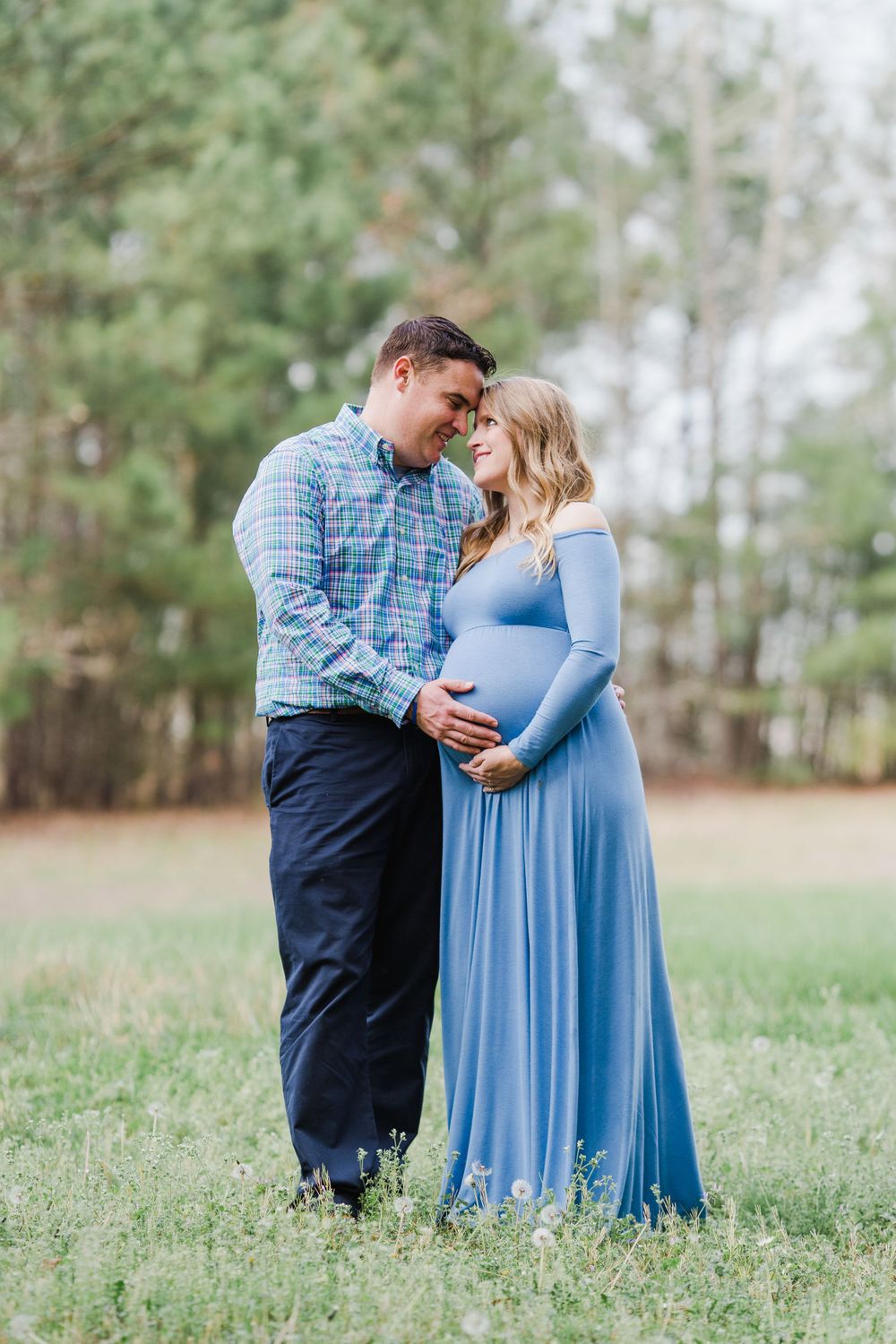 A maternity photo of a couple sharing an intimate moment in a grassy field with pine trees.