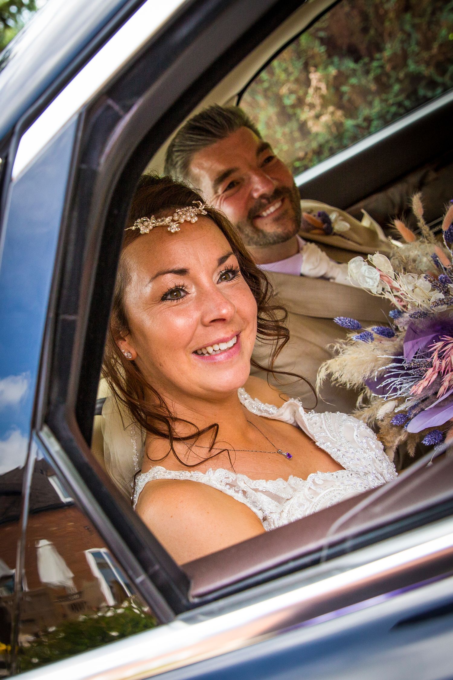 Bride with floral bouquet smiles joyfully from inside wedding car.