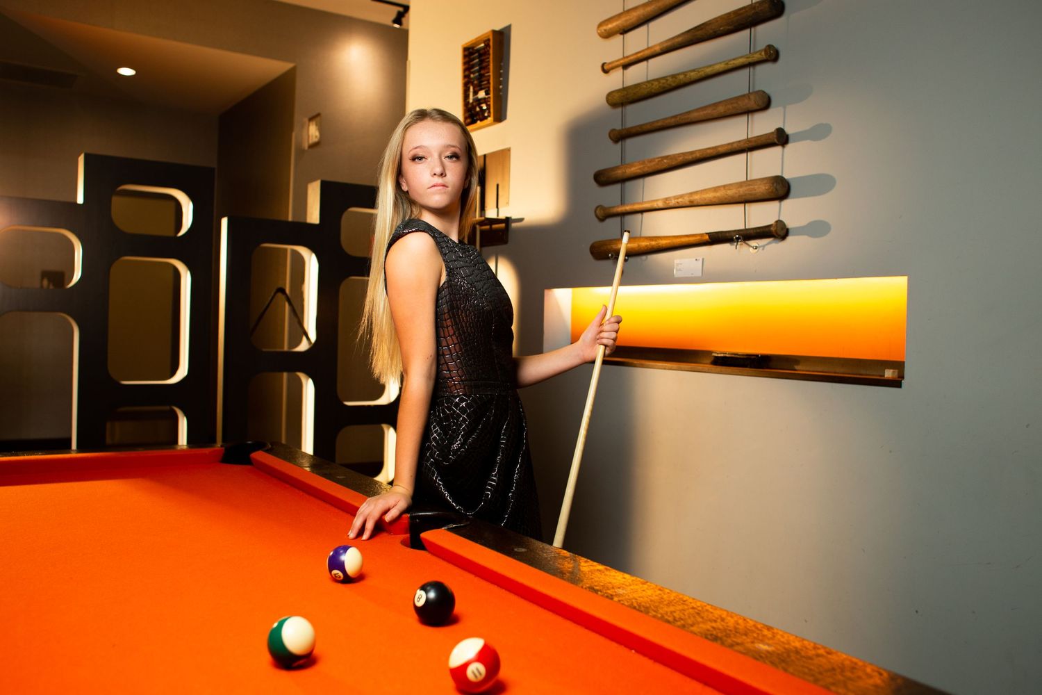 Woman in black dress poses next to orange pool table in dimly lit room.