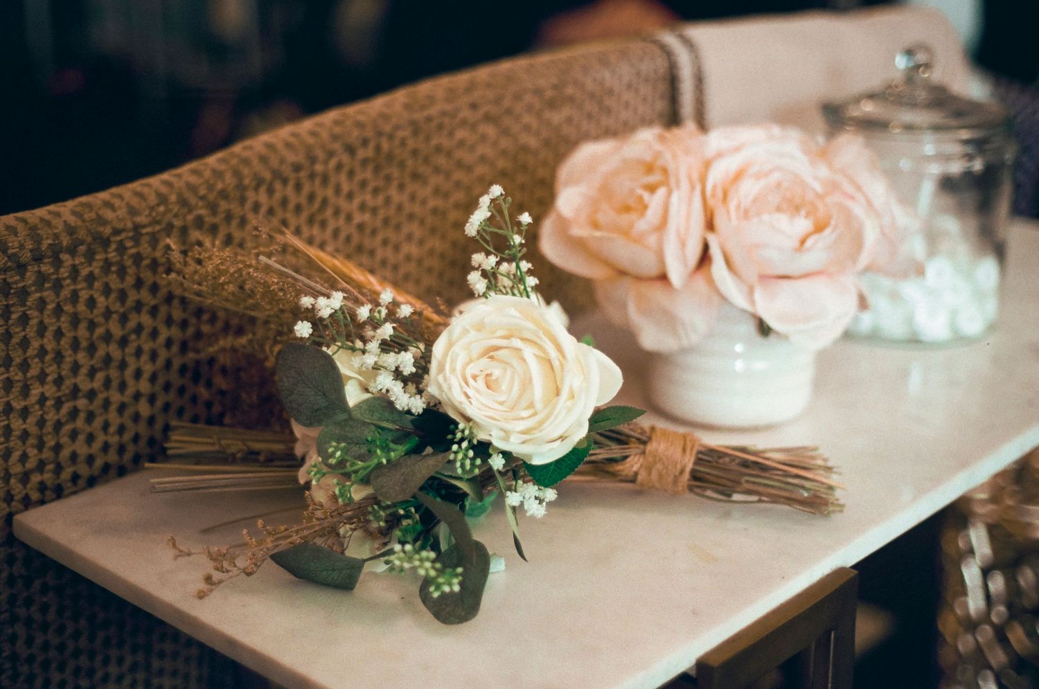 Delicate arrangement of pale pink and white roses with baby's breath on woven surface.