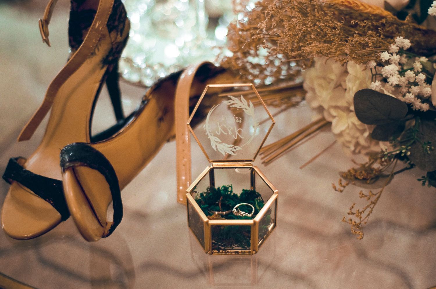 Gold hexagonal ring box with wedding rings displayed alongside floral arrangements and bridal shoes.