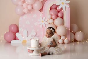 Baby girl sitting with her birthday cake wearing a daisy dress. Background setup is light pink balloons and arches with oversized daisies.