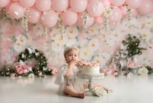 1st birthday baby girl wearing a ruffled white dress sitting with her cake. Backdrop is a light pink floral pattern and pink balloon garland