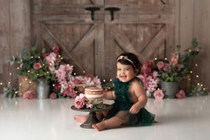 Happy baby girl wearing dark green sitting with her cake in front of a rustic barn wood backdrop accented with pink and green flowers.