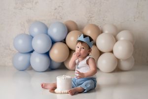 Baby boy sitting and feeding himself a bite of cake, he is wearing a light blue fabric crown and matching outfit, with tan and blue balloons