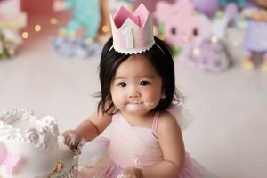 Closeup of a baby girl with cake on her face, she has on a pastel rainbow crown and dress.