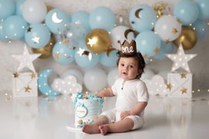 Happy baby boy sitting with his birthday cake in front of a twinkle star themed setup with balloons and stars in light blue, white and gold.