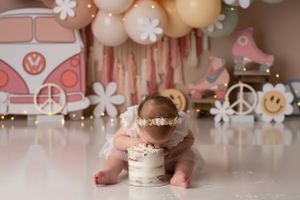 Baby girl with her face in her cake, the setup is a retro seventies theme with a VW bus and peace signs in muted rainbow colors.