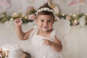 Closeup of a smiling birthday girl with cake on her face, she is wearing a white dress with white wings and flowers in the background.