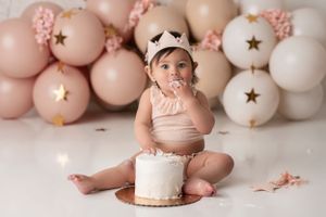 Baby girl feeding herself cake wearing a light peach outfit and crown, sitting with her cake in front of balloons in peach, tan and blush.