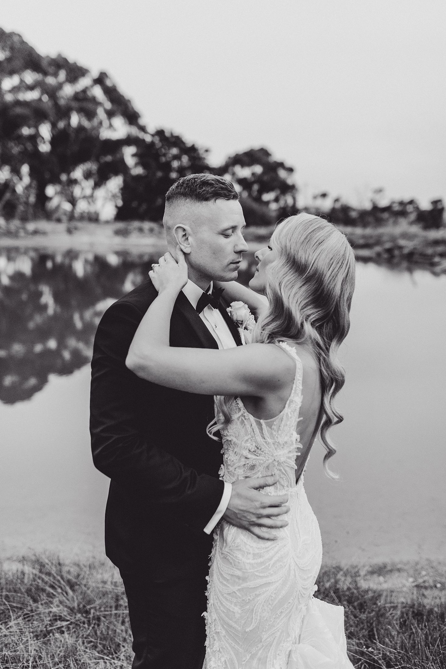 An intimate black and white portrait of a couple embracing on their wedding day by a lake.