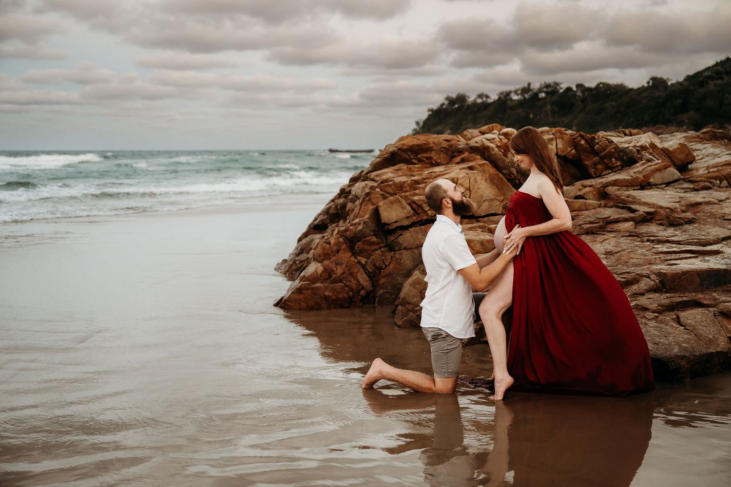Couple in formal attire poses romantically on rocky beach at sunset.