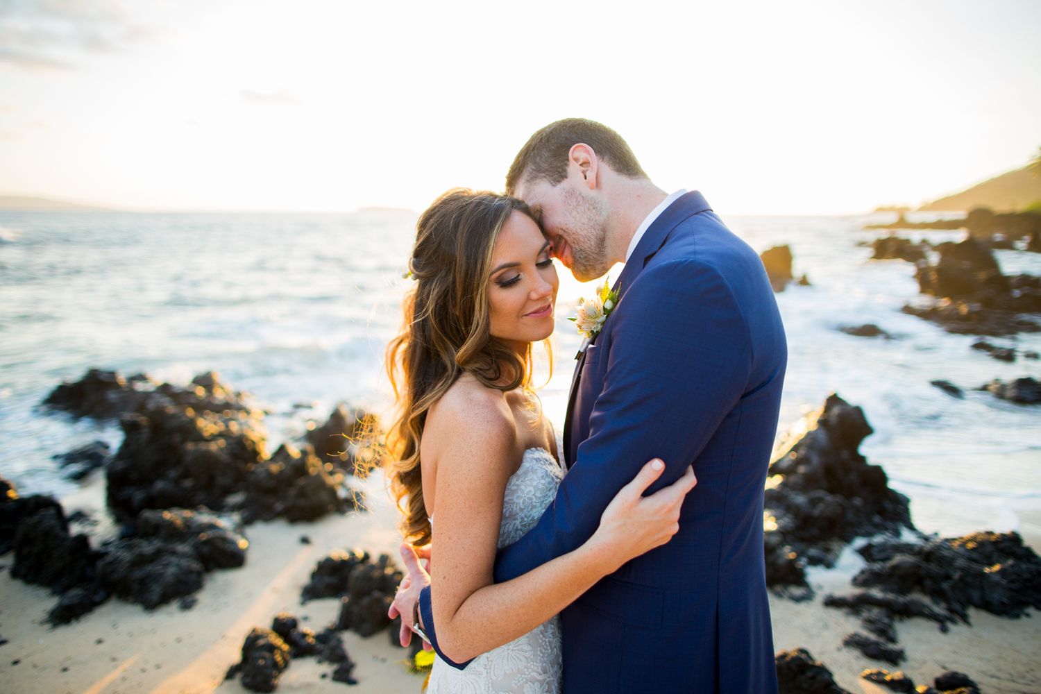 A couple shares an intimate moment on a rocky beach at sunset with crashing waves in the background.