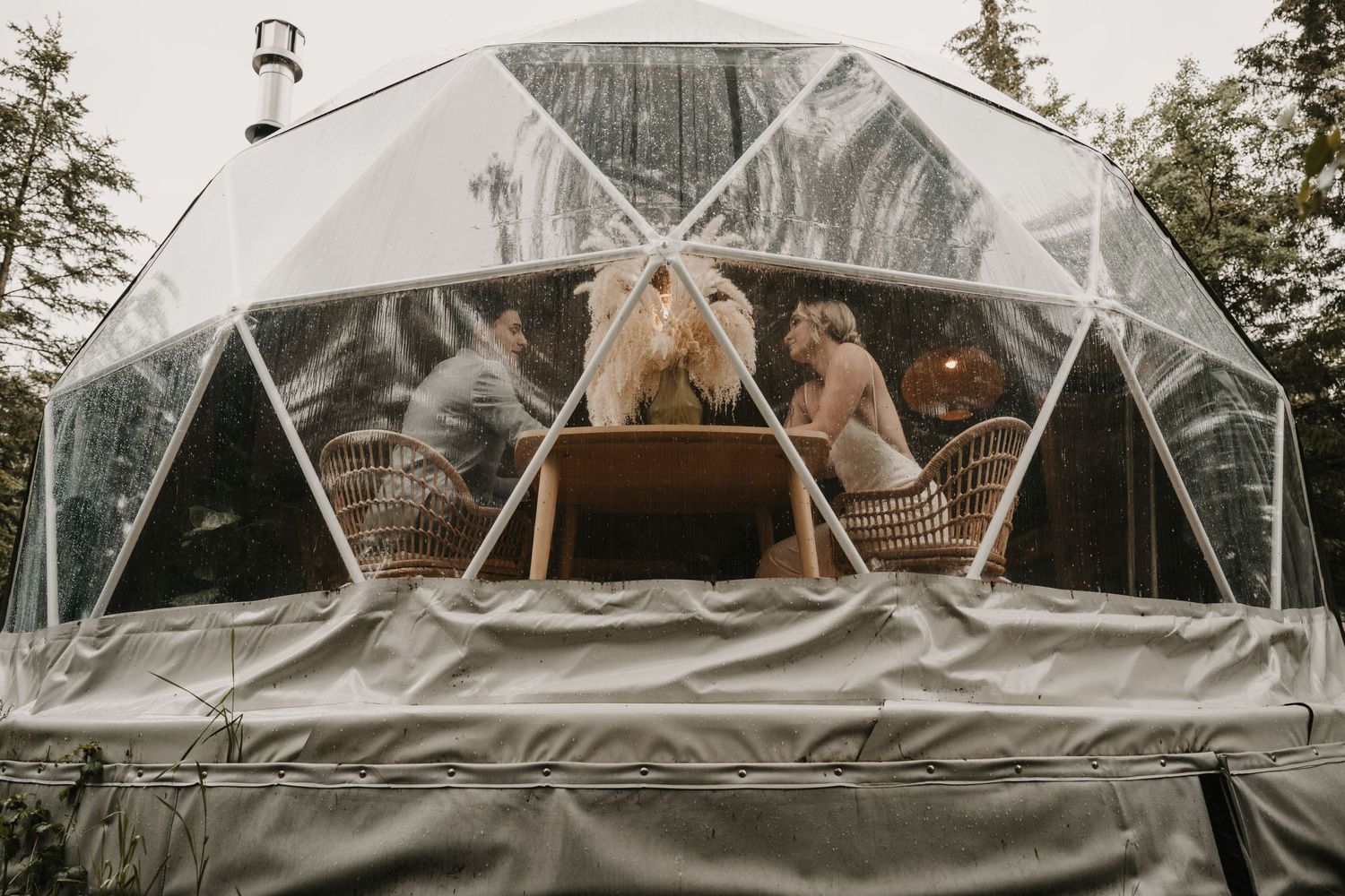 A cozy scene inside a clear geodesic dome tent with people relaxing on cushions and blankets in the wilderness.