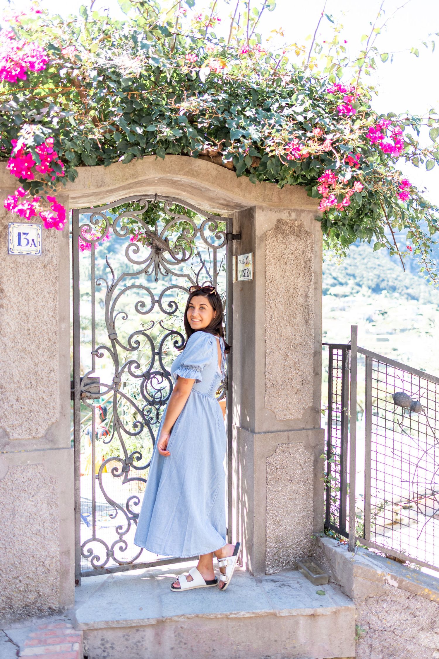 Wedding photographer in light blue dress stands by an ornate wrought iron gate under an arch with blooming pink bougainvillea flowers.