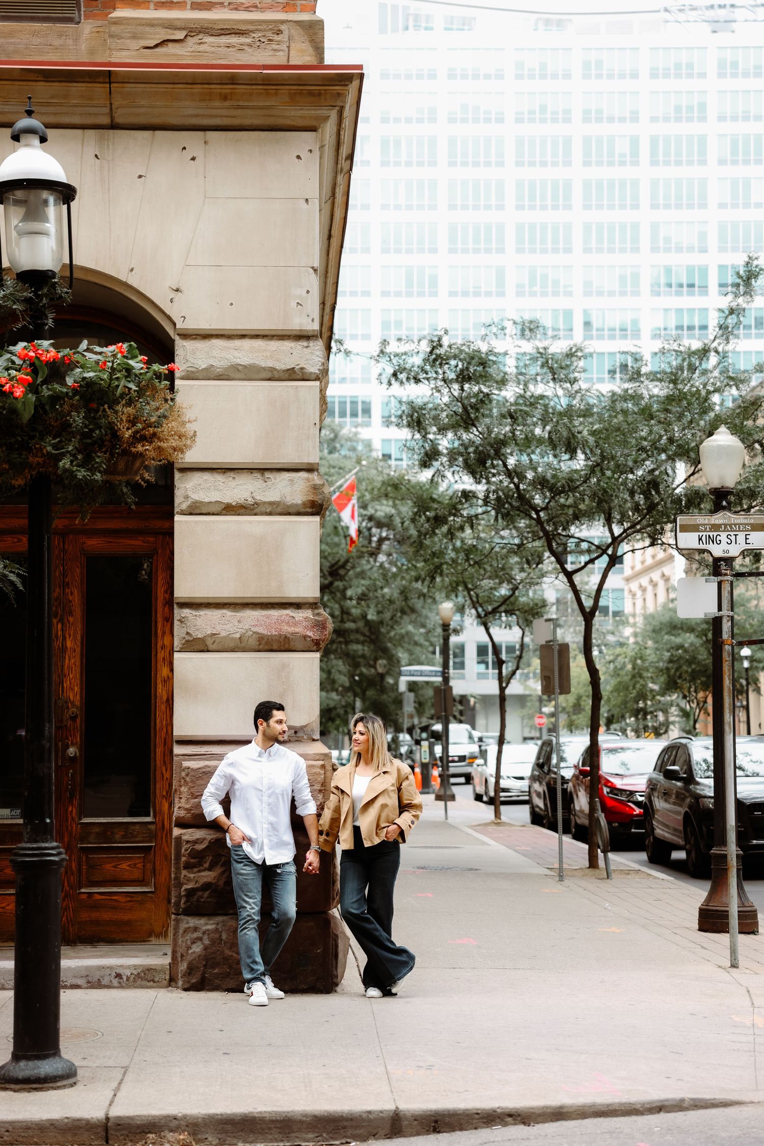 Stylish couple strolls down a city sidewalk past historic architecture and flowering planters.