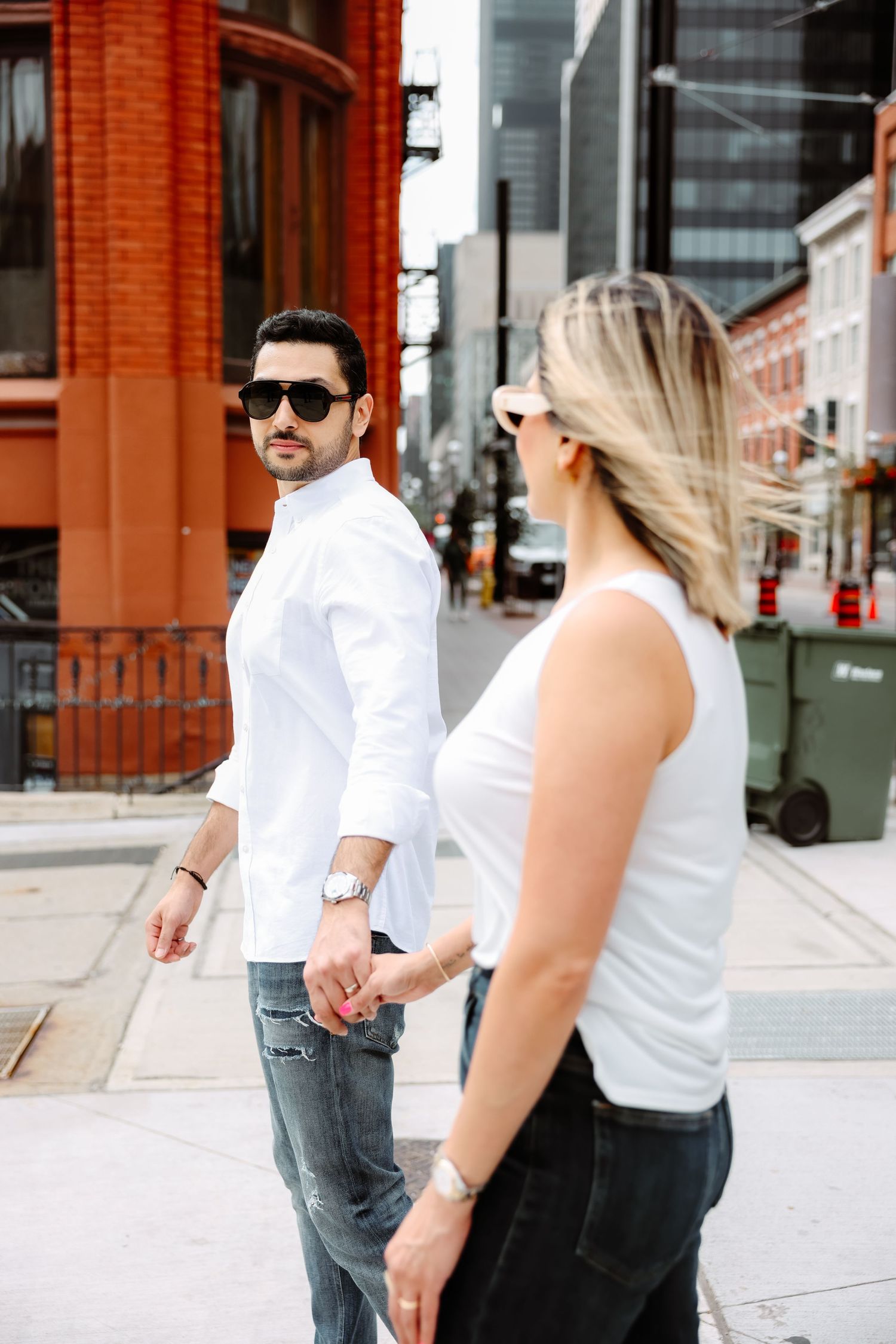 Couple in coordinated white outfits walking together on a city street with red brick buildings in the background.