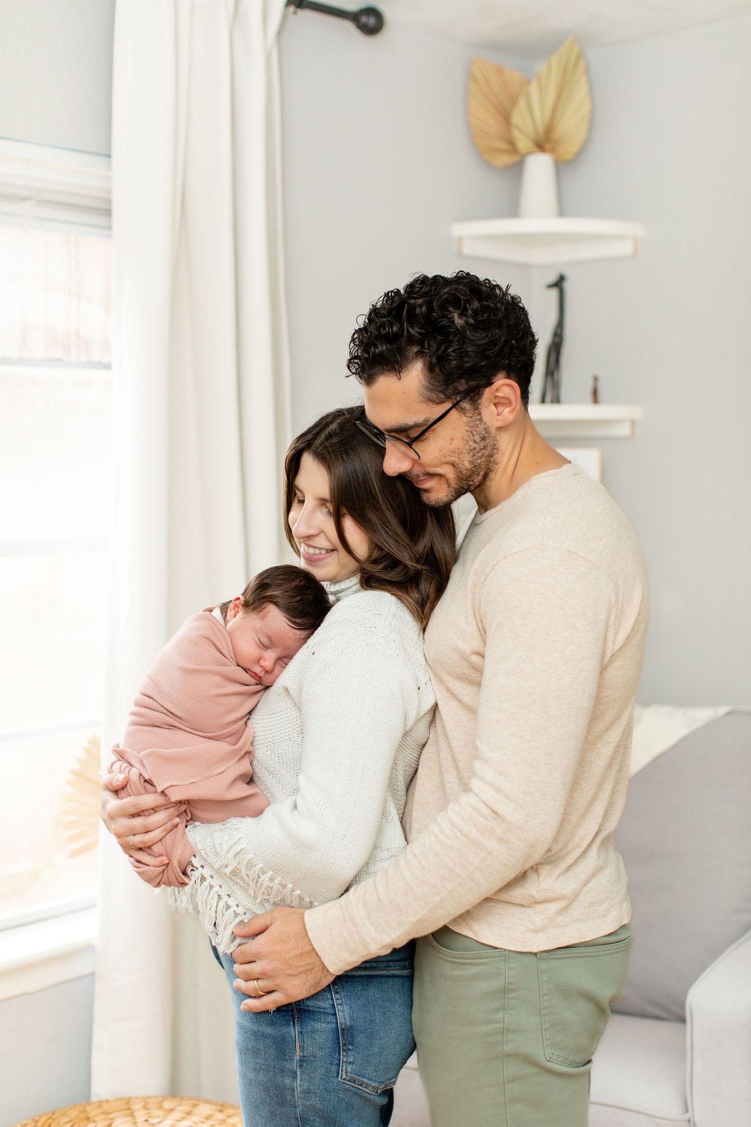 A couple embraces while holding their newborn baby in a cozy living room setting.
