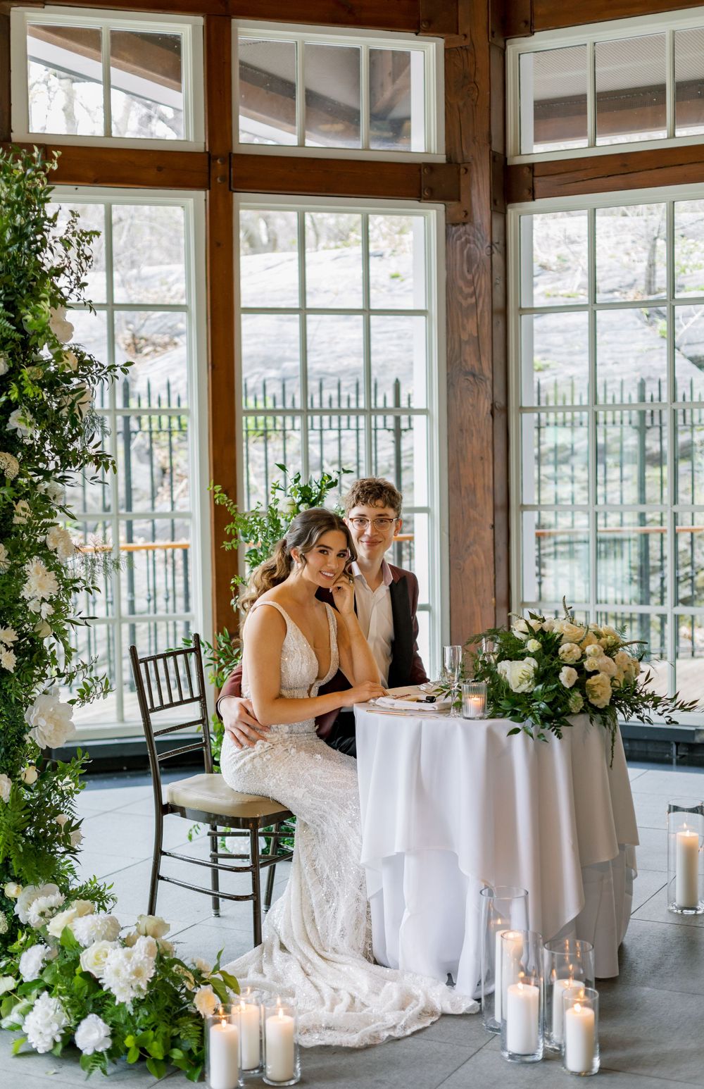 Elegant wedding couple seated at a decorated table in a sunlit conservatory with candles and flowers.