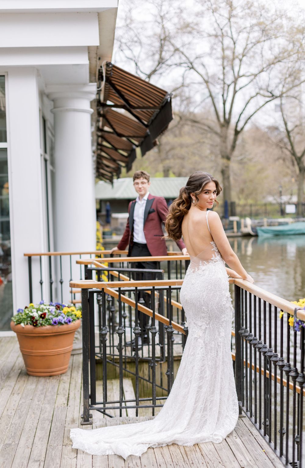 Bride in fitted lace gown poses on waterfront deck while groom stands in background.