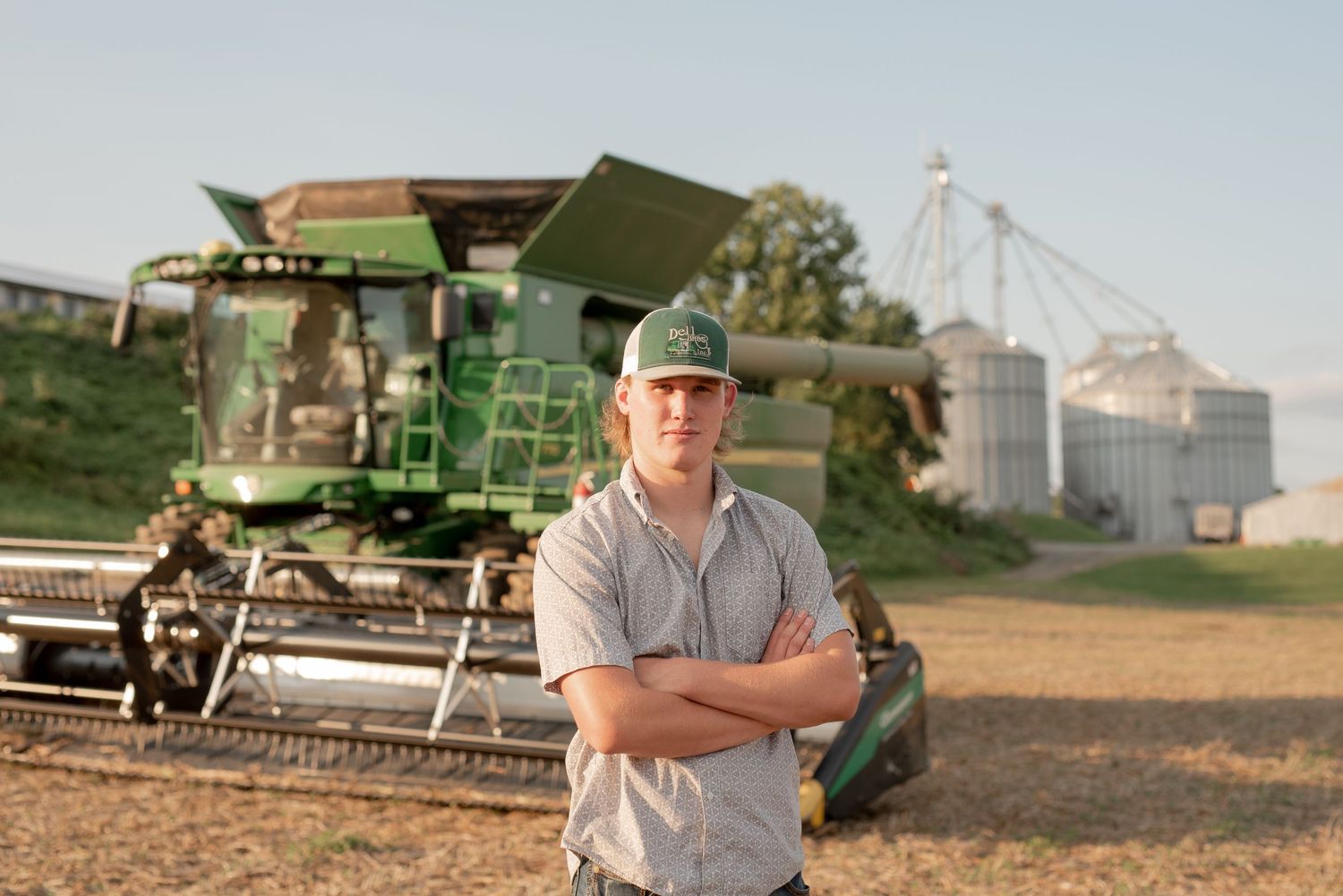 Farmer standing in front of green combine harvester and grain silos at sunset.