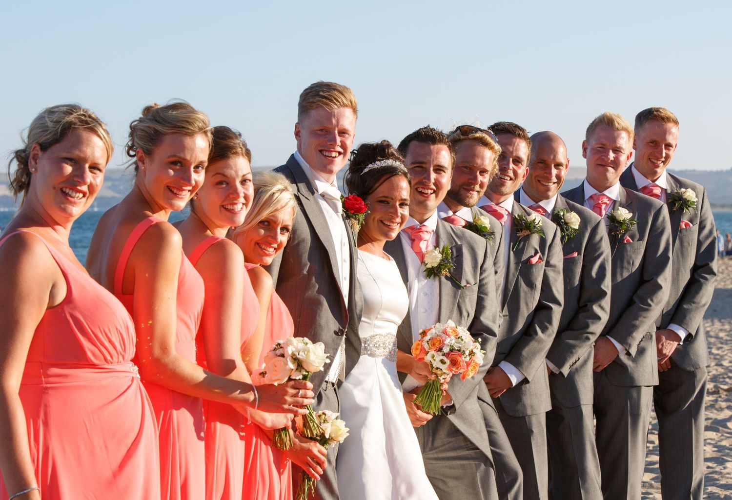 Wedding party lineup with bridesmaids in coral dresses and groomsmen in gray suits outdoors.