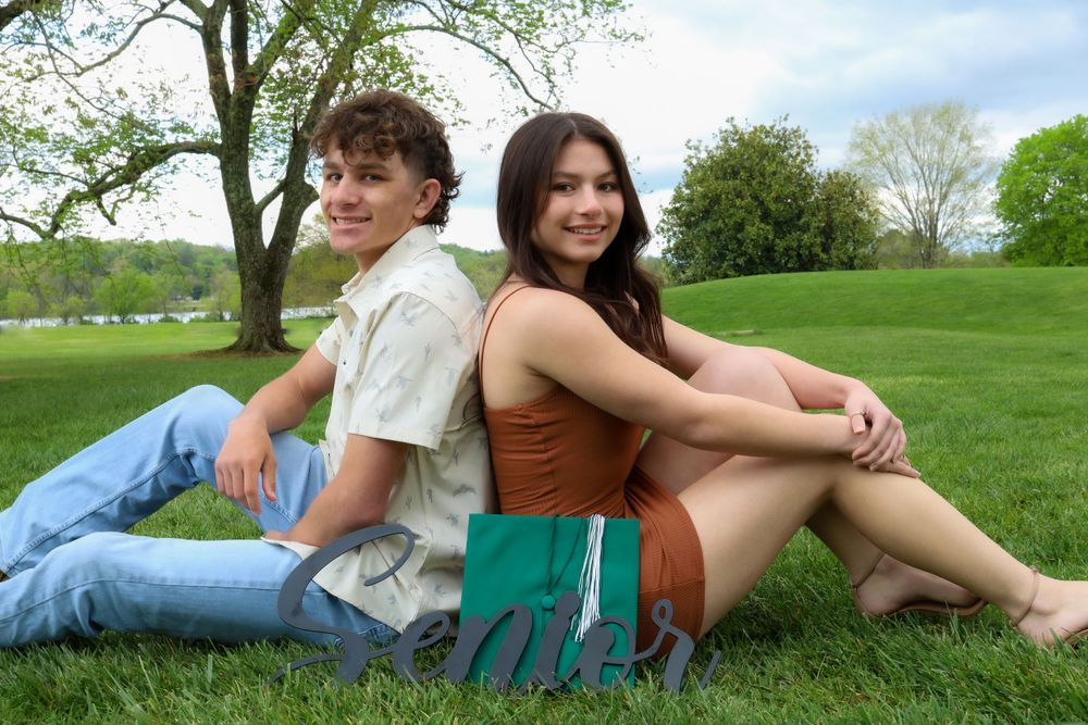 Two people sitting back-to-back on green grass in a park on a sunny day.