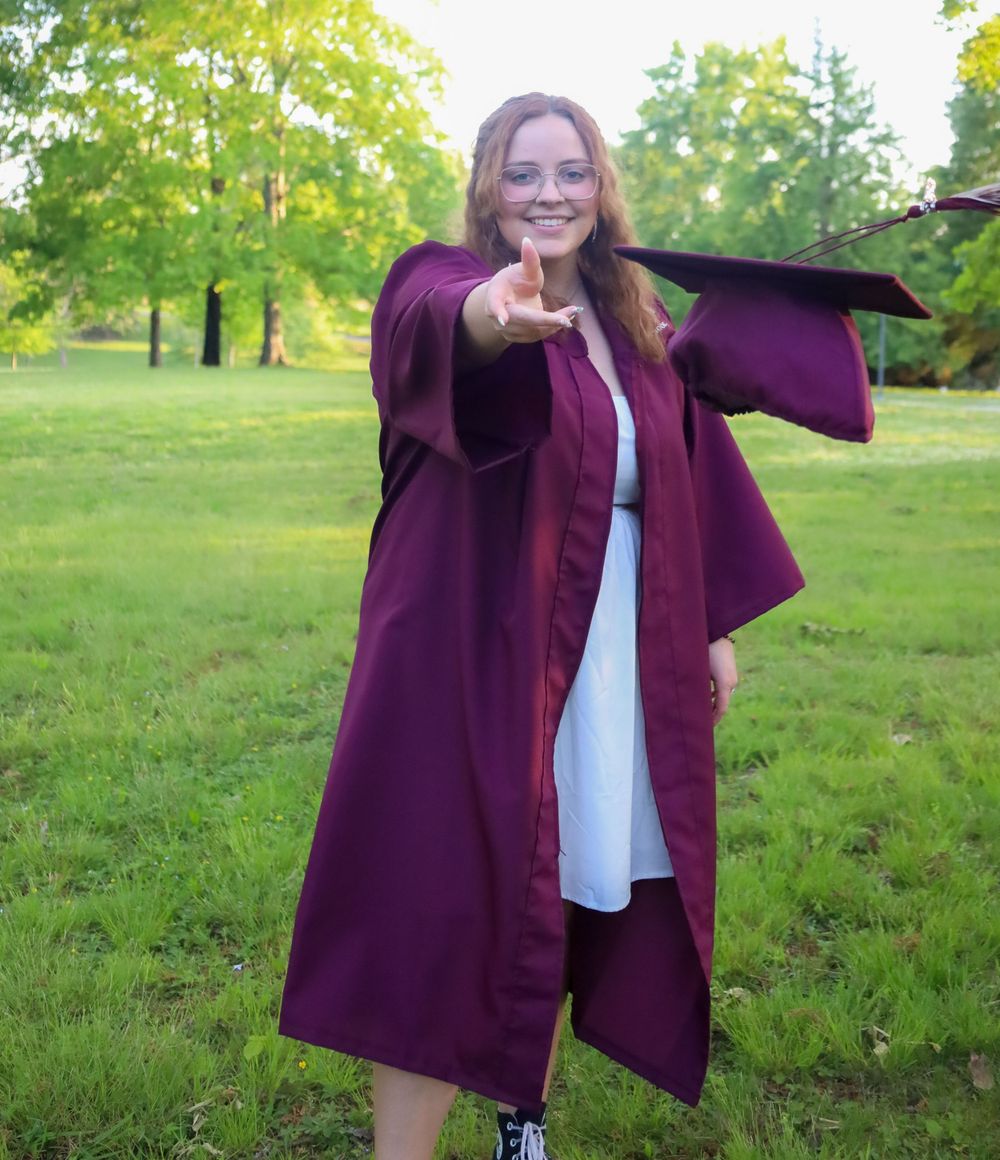 Graduate in maroon gown poses outdoors in a sunny grassy field.