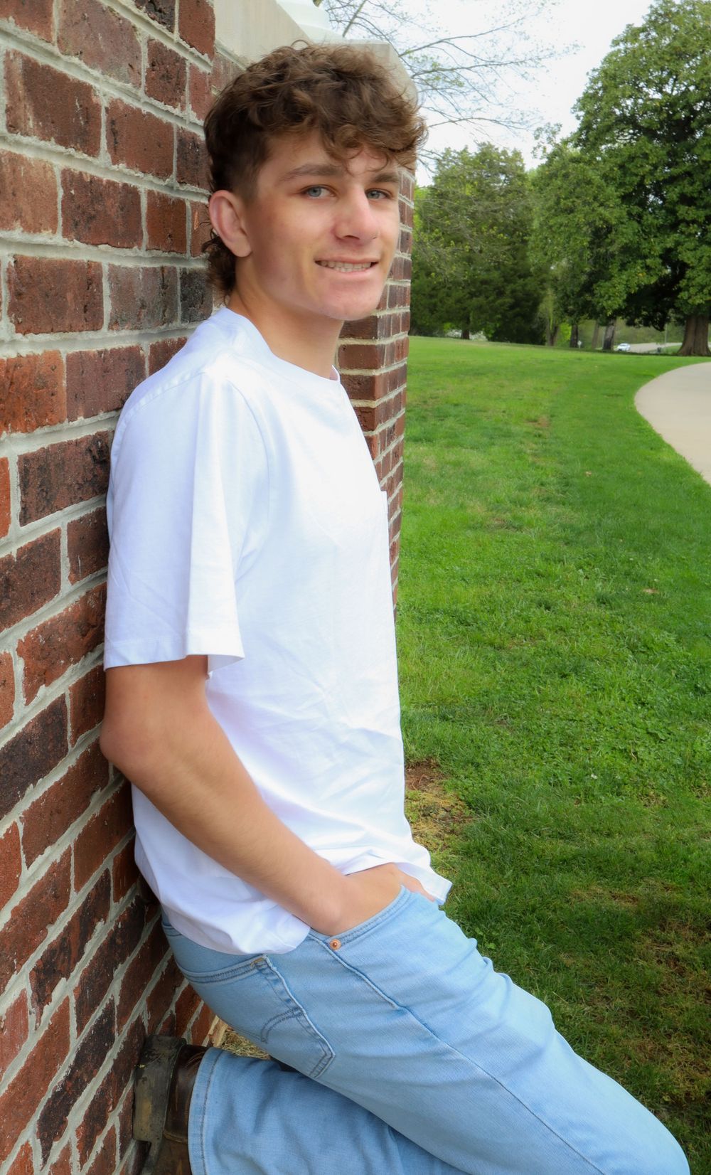 Person in white t-shirt leaning against brick wall with green lawn in background.