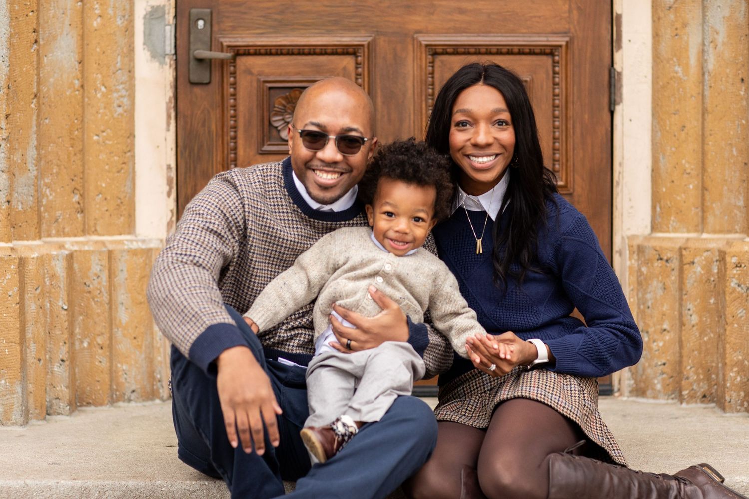 A smiling family sits together on stone steps in front of a wooden door.