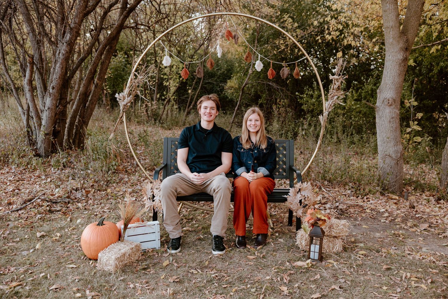 A couple sits on a bench with pumpkins and a dog under a decorated arch with string lights.