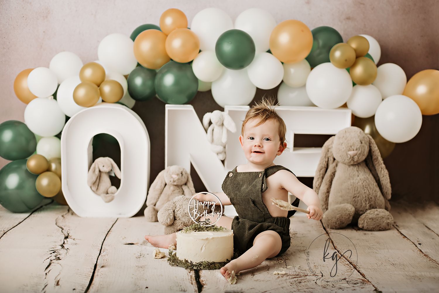 First birthday setup with green and gold balloons, stuffed animals, and cake.