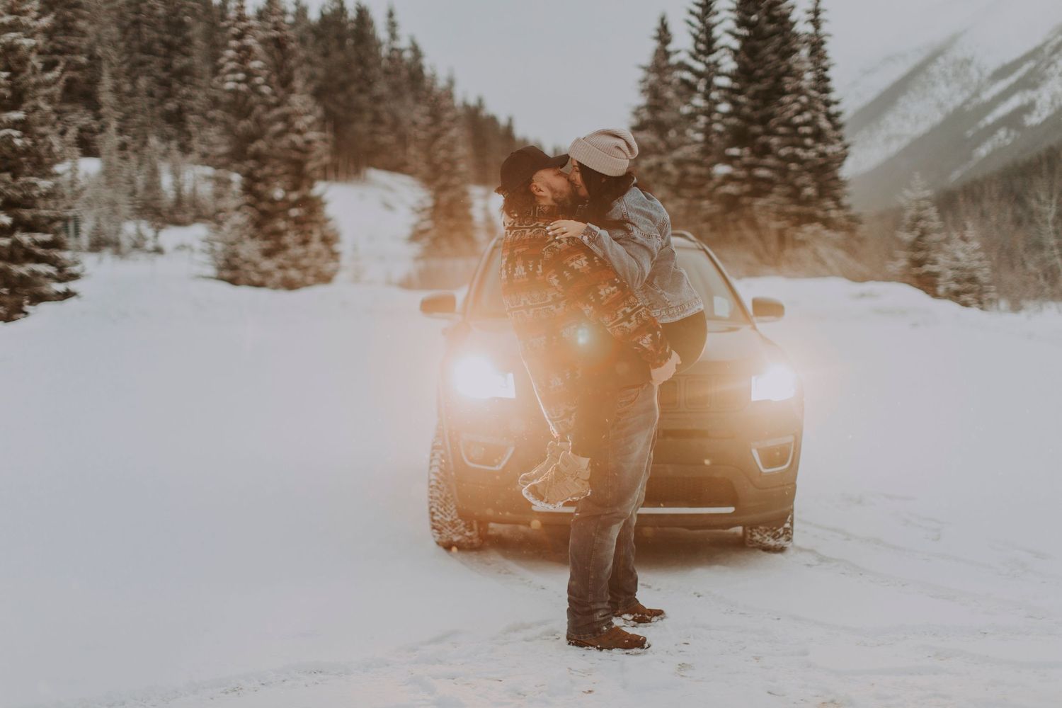 A car with bright headlights illuminates a snowy winter mountain road scene.