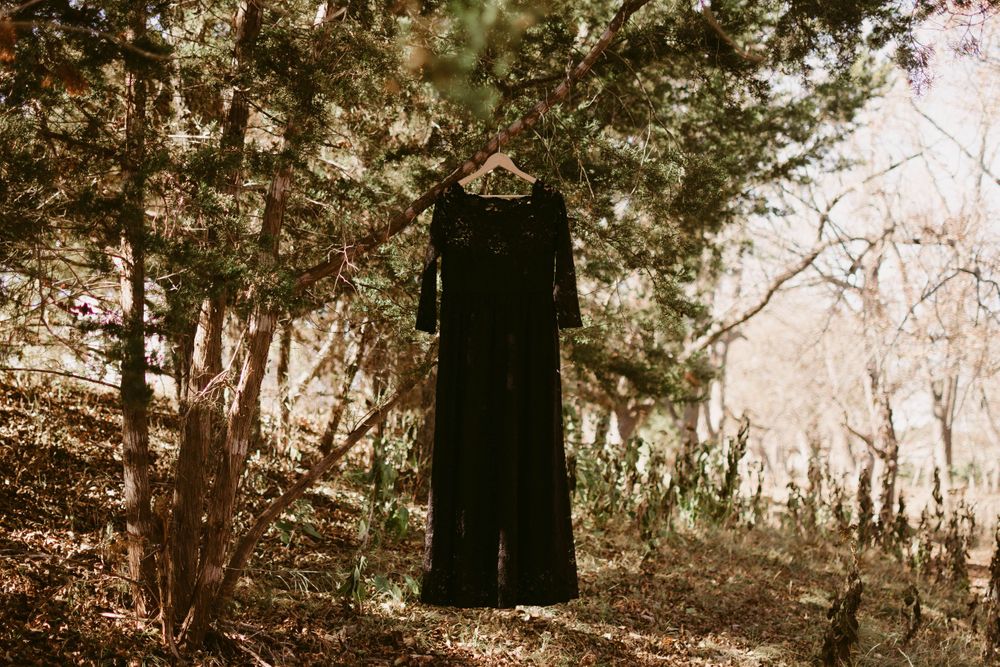 Long black dress hanging on a wooden hanger among trees in a misty forest setting.
